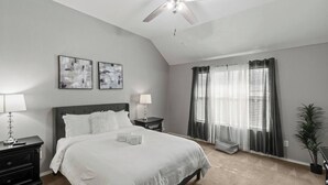 Une chambre à coucher avec un lit, deux tables de chevet, un ventilateur de plafond, une fenêtre avec des rideaux et une plante.