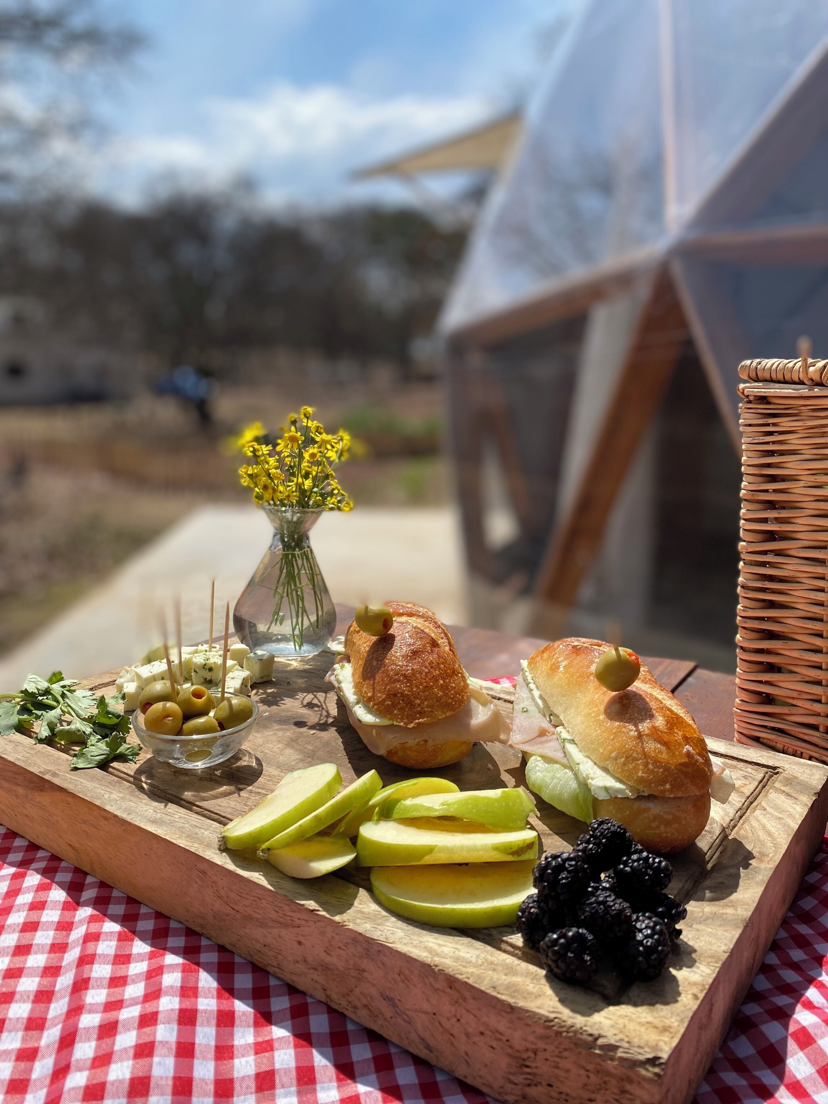 A wooden board with food items, a wicker basket, and a checkered tablecloth.