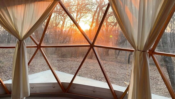 Una habitación con diseño geométrico en el techo, ventanas amplias con cortinas y vistas a árboles y un atardecer.