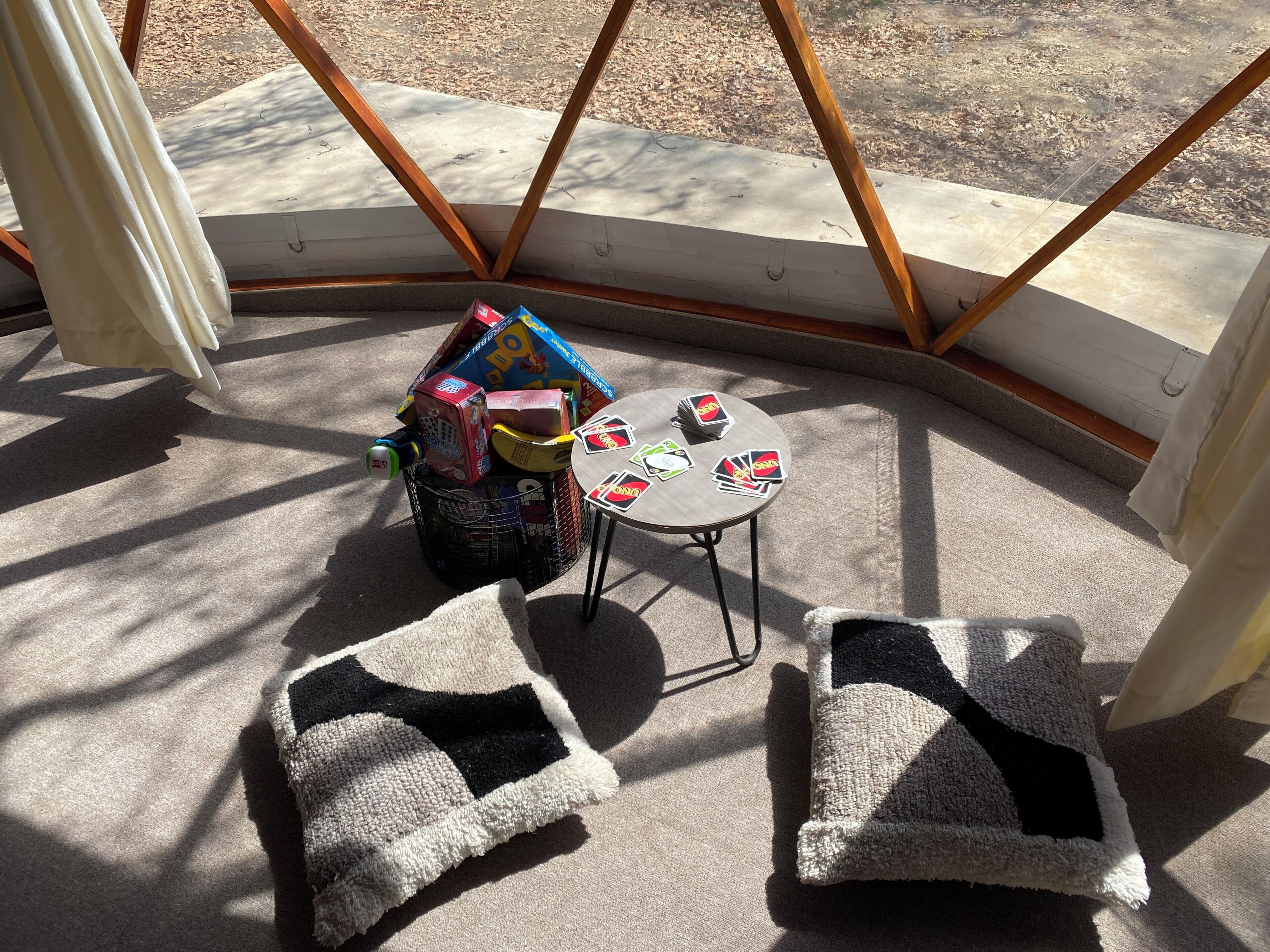 Una esquina de una habitación con una mesita, dos cojines, una bolsa de compras y un ventanal con vistas al exterior.