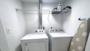 A laundry room with two white washing machines, a metal shelf above, and a beige curtain. - Welcome to Heidi's Nest! Renovated 2BR Condo in Bradenton Beach/Runaway Bay! (Bradenton Beach)