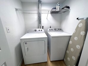 Ein Waschraum mit zwei weißen Waschmaschinen, einem Metallregal darüber und einem beige farbenen Vorhang.