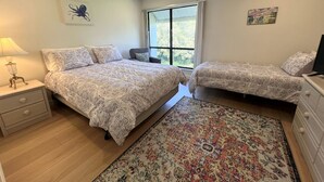 A bedroom with two beds, a nightstand, a dresser, a window, and a rug. - Welcome to Heidi's Nest! Renovated 2BR Condo in Bradenton Beach/Runaway Bay! (Bradenton Beach)