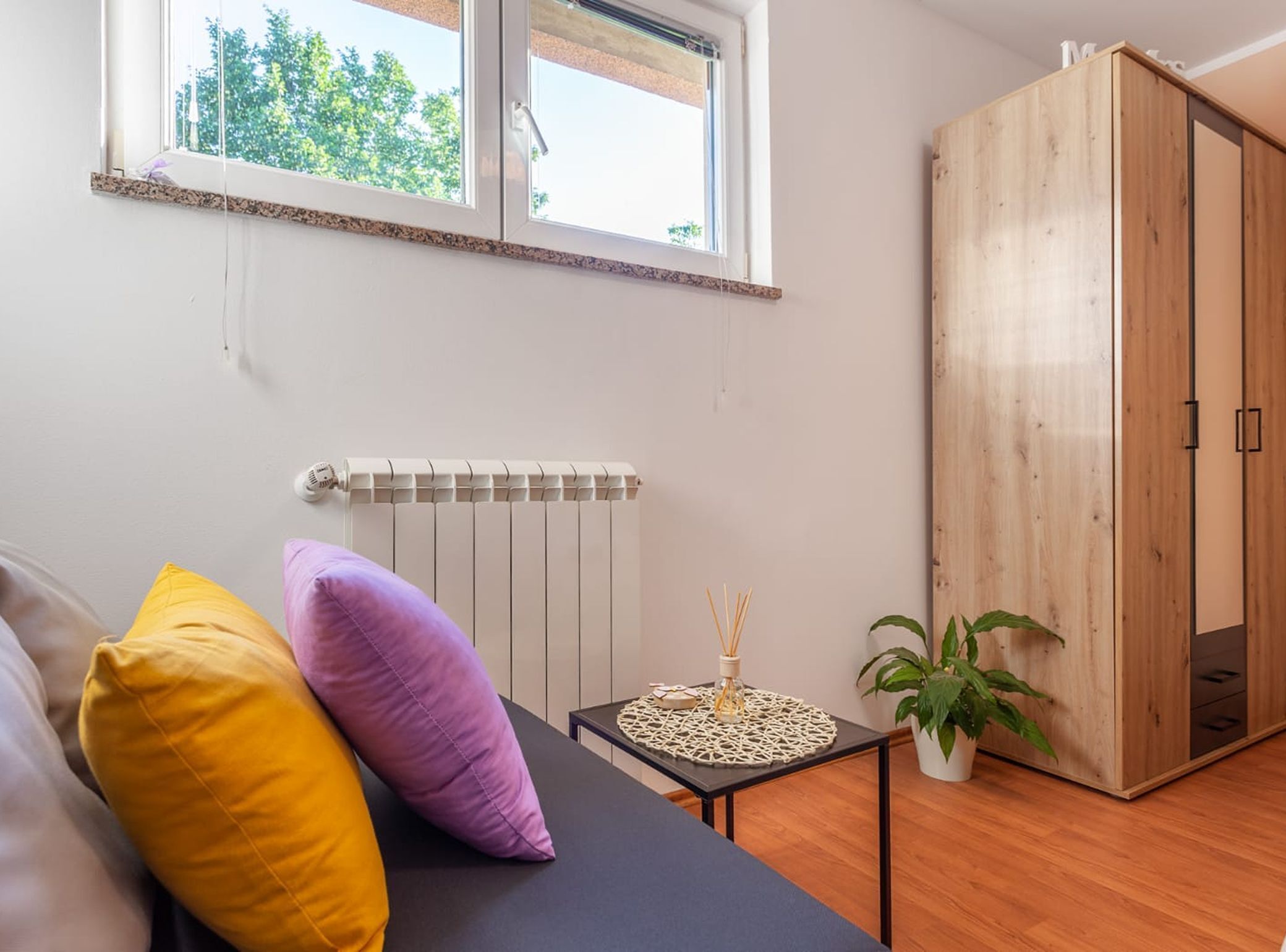 A room with a bed, a radiator, a window, a wardrobe, and a small table with a plant.