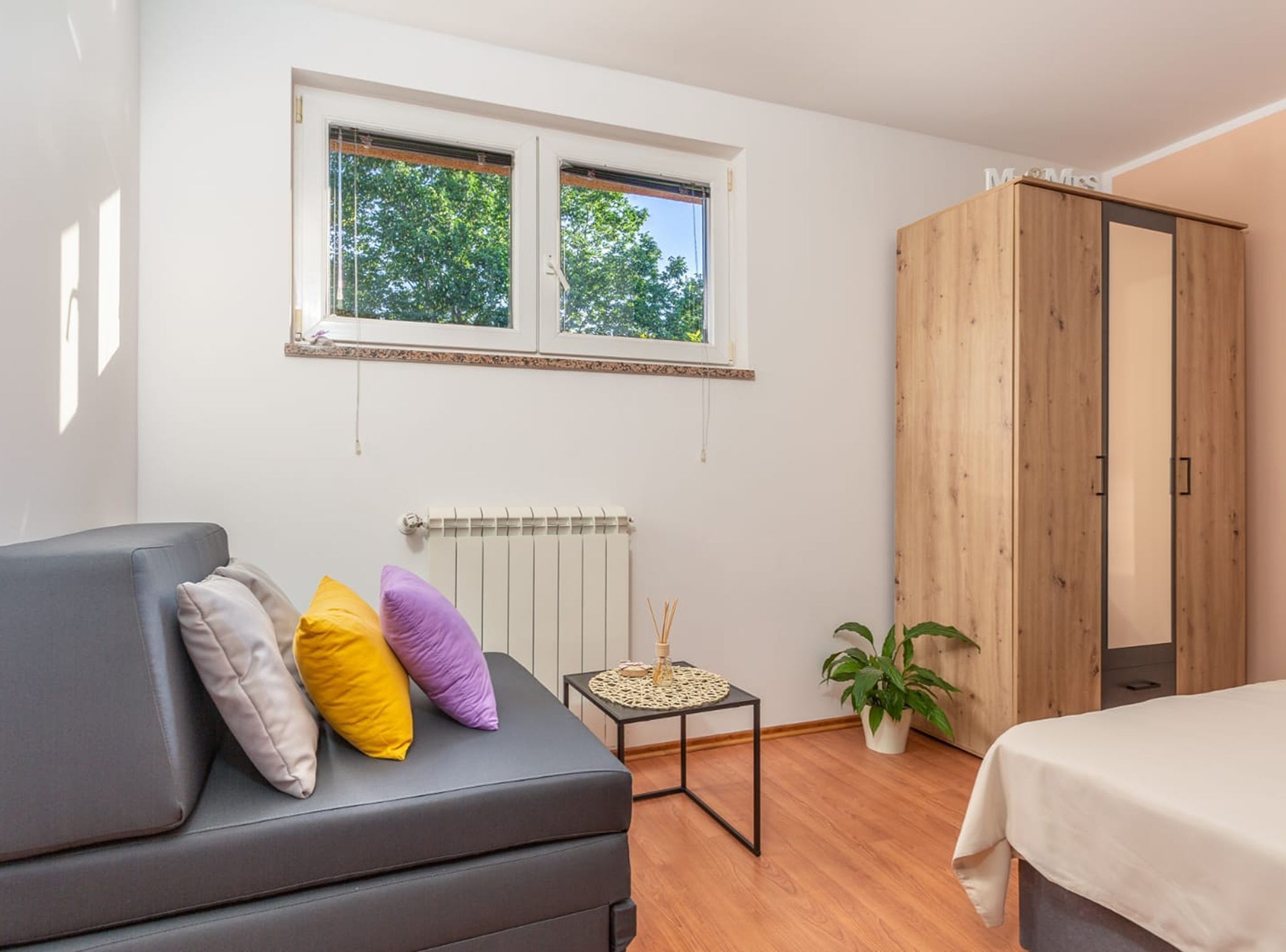 Et soveværelse med en grå sofa, et træskab, et lille bord med en plante og en radiator.