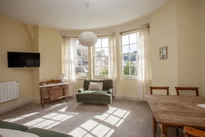 A room with a green armchair, a wooden table, a television mounted on the wall, and large windows with curtains. - Hidden Georgian Apartment - Central Bath (Bath)