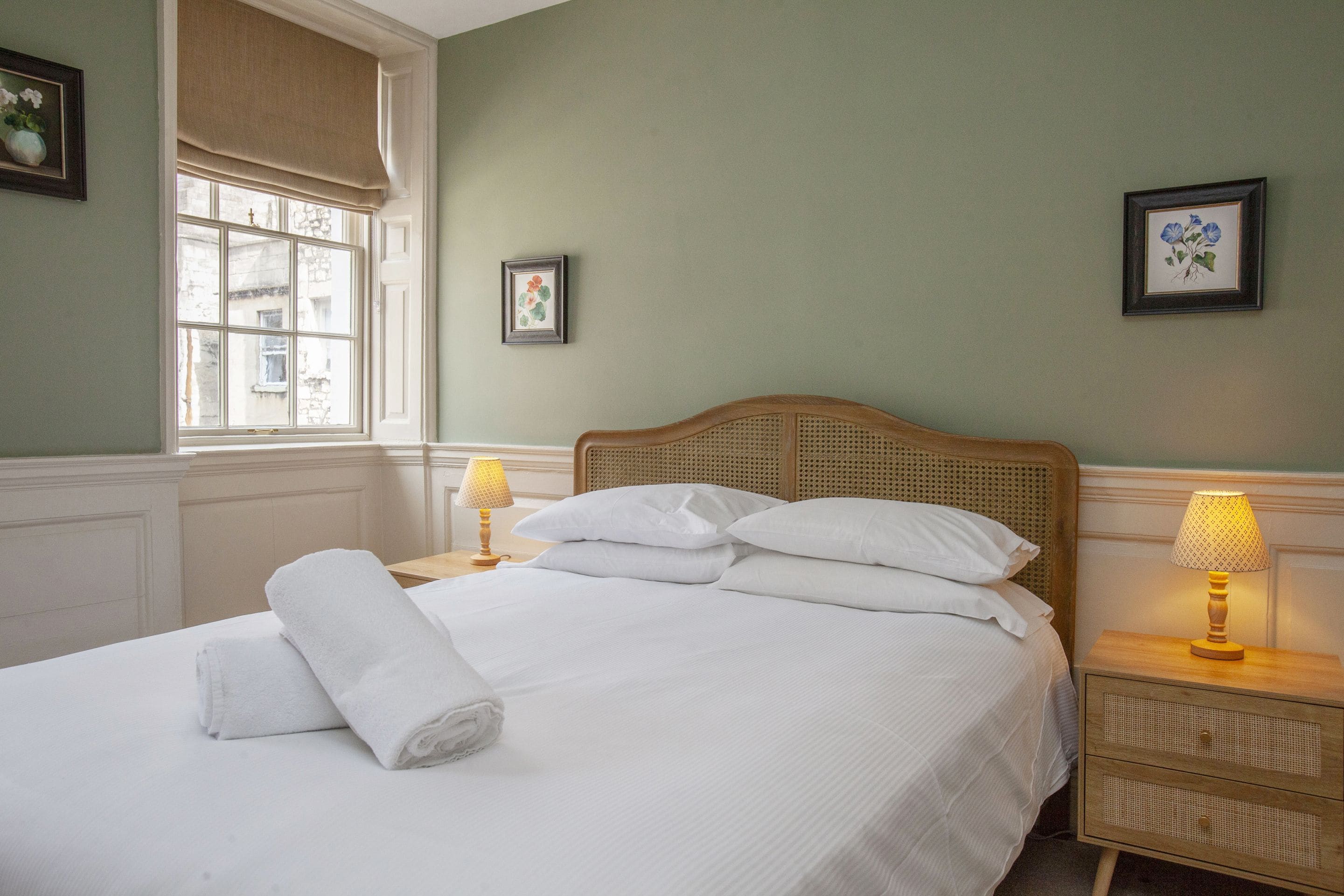 A neatly made bed with white linens and pillows, a window with a view of buildings, and two bedside lamps on wooden nightstands.