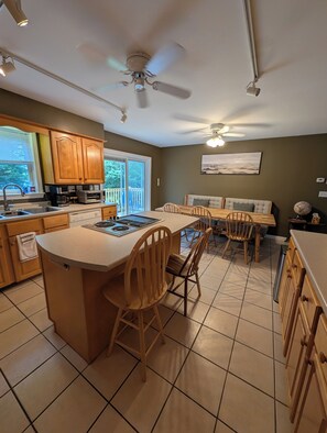 A kitchen with wooden cabinets, a central island with chairs, a ceiling fan, and a dining area with a view of the outdoors. - Lakeside Haven on Whites Pond (Palmyra)