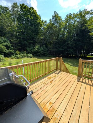 Eine Holzterrasse mit Grill und Spüle, umgeben von üppiger, grüner Landschaft und Bäumen.
