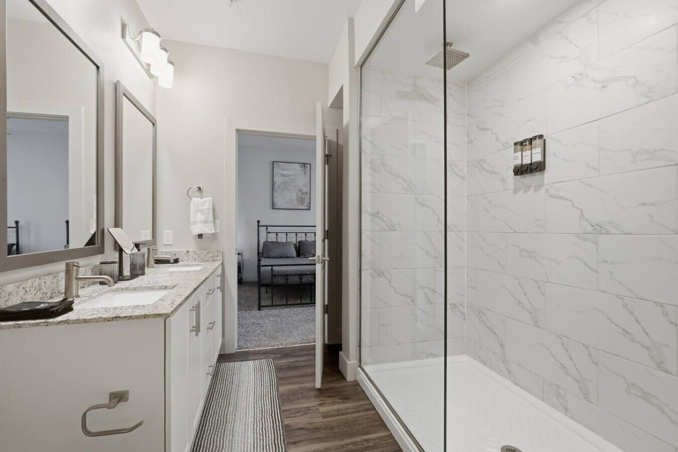A modern bathroom with a glass shower, marble walls, and a bed in the adjacent room.