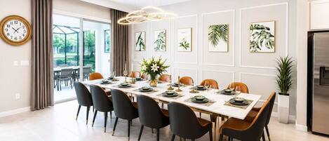 Une salle à manger moderne avec une longue table, des chaises, une horloge et des œuvres d’art encadrées accrochées au mur.