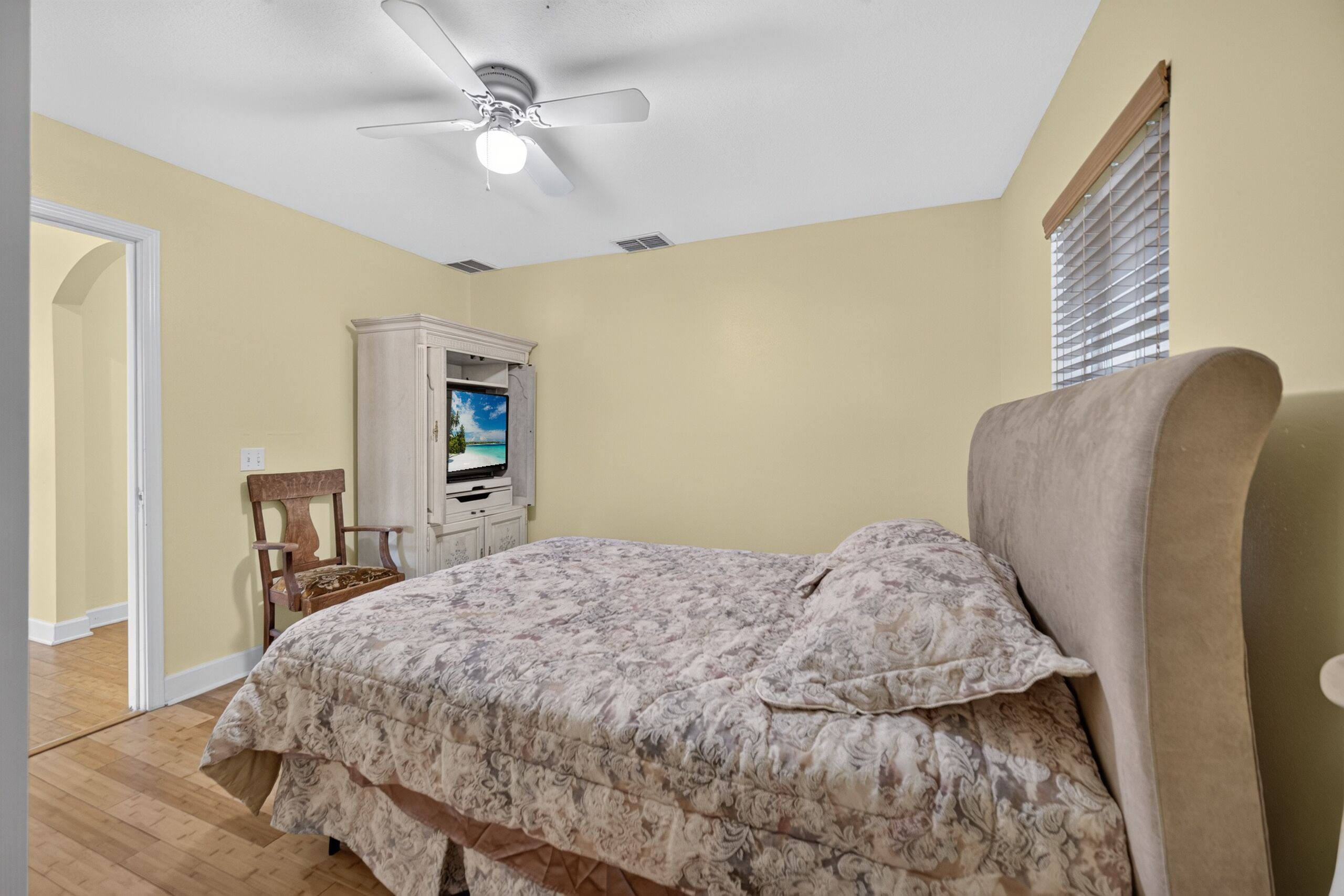 A bedroom with a bed, a ceiling fan, a television, and a window with blinds.
