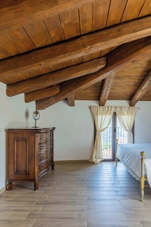 Ein Schlafzimmer mit Holzbalkendecke, einer Holzkommode, einem Bett mit Metallgestell und einem Fenster mit Vorhängen.