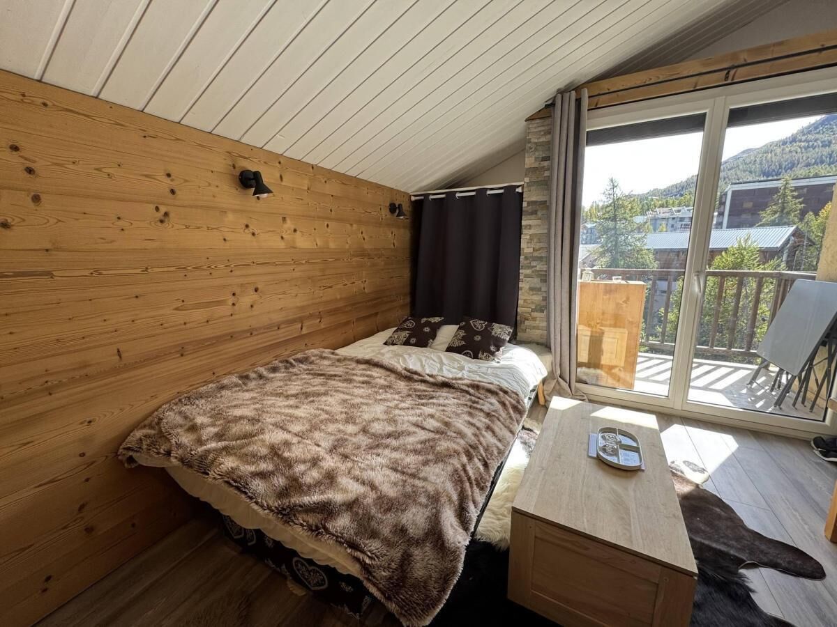 Una camera da letto con soffitto in legno, un letto con coperta marrone, un comodino in legno e una finestra vista edifici e alberi.
