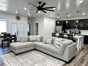 A modern living room with a sectional sofa, a dining area, and a kitchen with dark cabinets and stainless steel appliances.