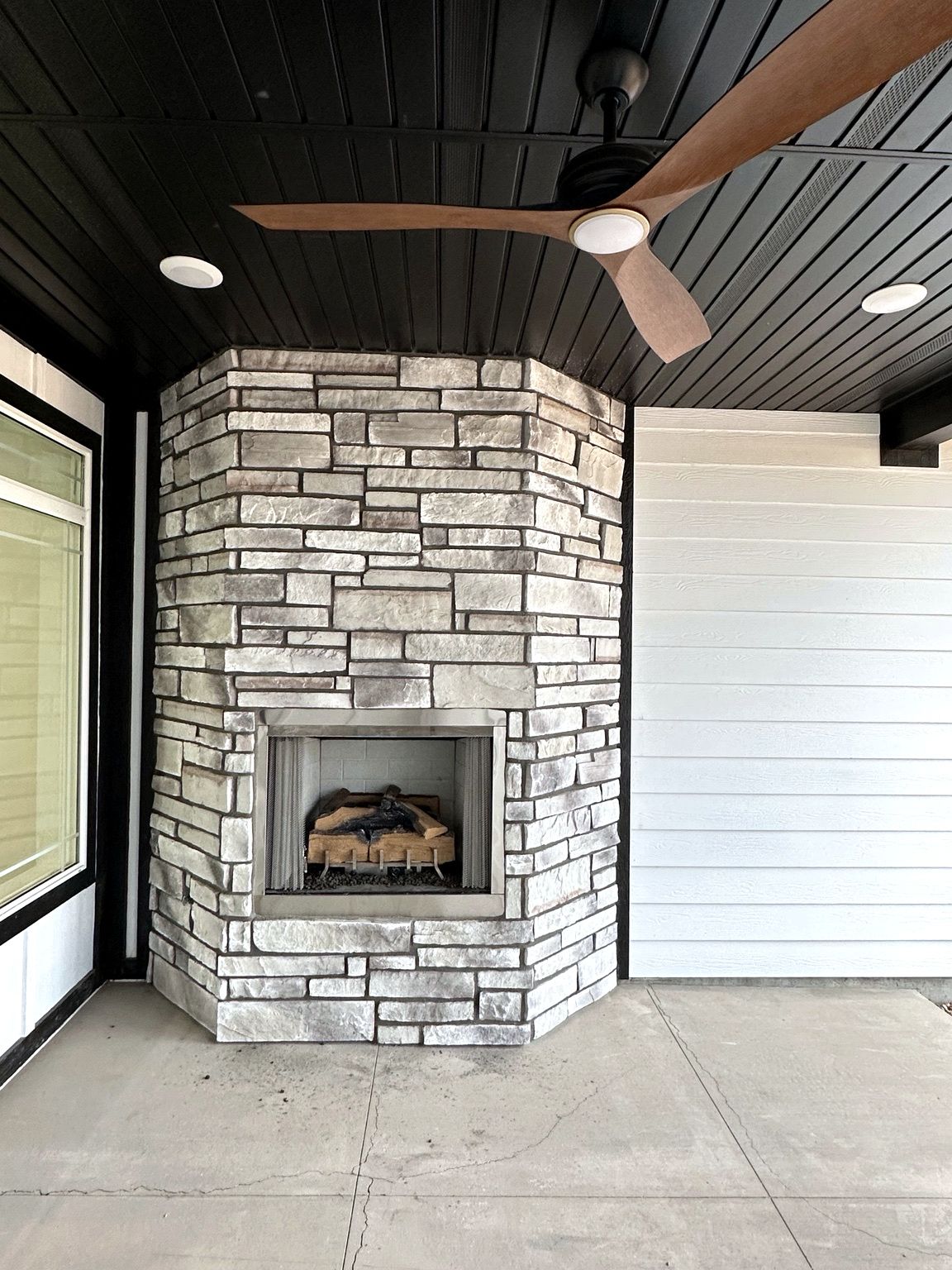 Une cheminée en pierre, surmontée d’un ventilateur de plafond, est installée dans un coin, au-dessus d’un plancher en béton.