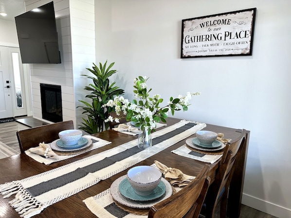 A dining area with a wooden table set for four, surrounded by potted plants and a welcome sign.