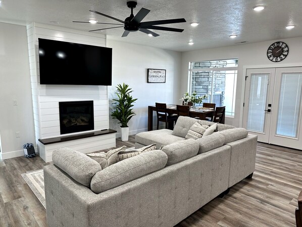 A modern living room with a sectional sofa, a flat-screen TV mounted above a fireplace, a ceiling fan, and a dining area with a table and chairs.