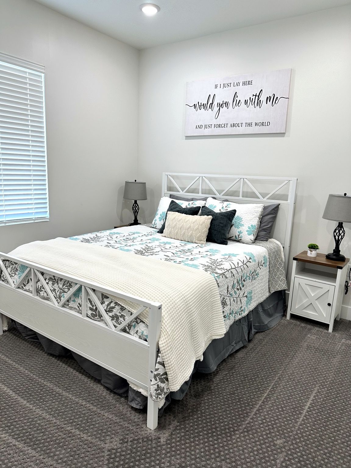 A bedroom with a bed, two bedside tables, a wall-mounted sign, and a window with blinds.