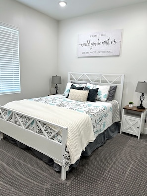 A bedroom with a bed, two bedside tables, a wall-mounted sign, and a window with blinds.