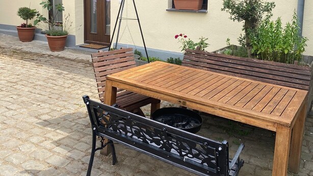 A wooden table and bench set outside a building with a window and door, potted plants, and a clear sky. - An Eifel idyll at the Nürburgring: A spacious oasis of comfort with plenty of room (Herresbach)