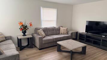 A modern living room with a grey sofa, a coffee table, a TV stand, and a window with blinds.