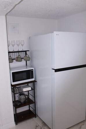 A compact kitchen with a white refrigerator, a microwave, and a shelf holding wine glasses and mugs. - Delightful 1-bedroom studio with AC in Port Richey (Port Richey)
