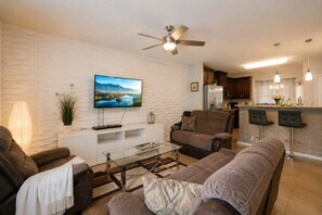 A modern living room with a flat-screen TV, a sofa, a coffee table, and a ceiling fan. - Meadow Manor - 10 Mins to Disney! - No Rear Neighbors, Covered Lanai (Kissimmee)