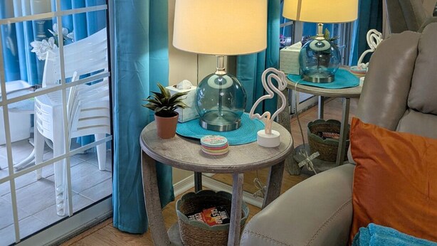 A room with a view through a glass door, a sofa, a table with a lamp, and a small plant. - Secrets Under the Palms - Luxury 1BR King Condo, Pond View Lanai (Lutz)