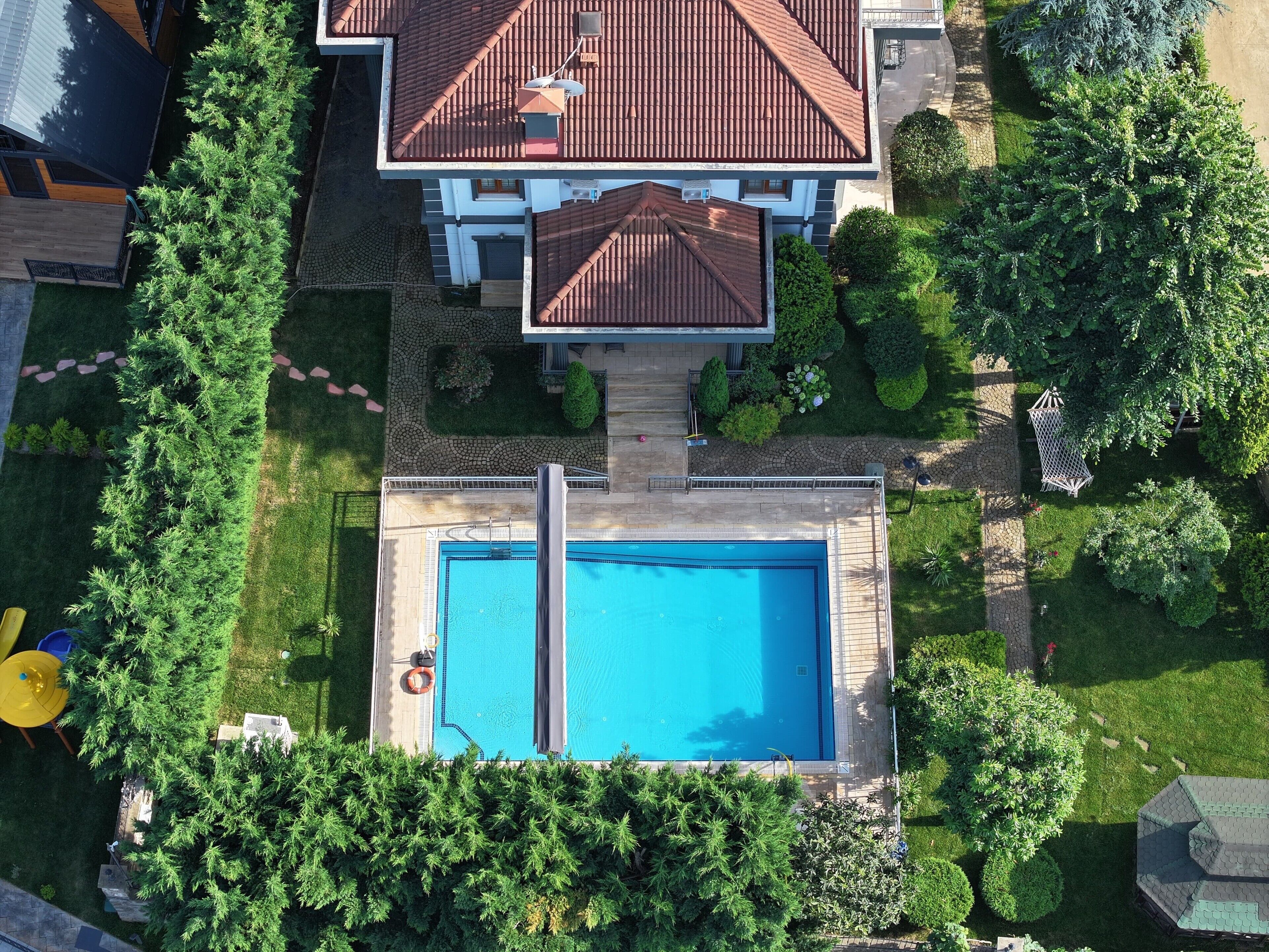 An aerial view of a residential property with a swimming pool, a two-story house with a red-tiled roof, and surrounding greenery.