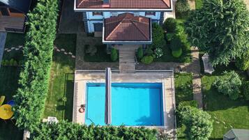 An aerial view of a residential property with a swimming pool, a two-story house with a red-tiled roof, and surrounding greenery.