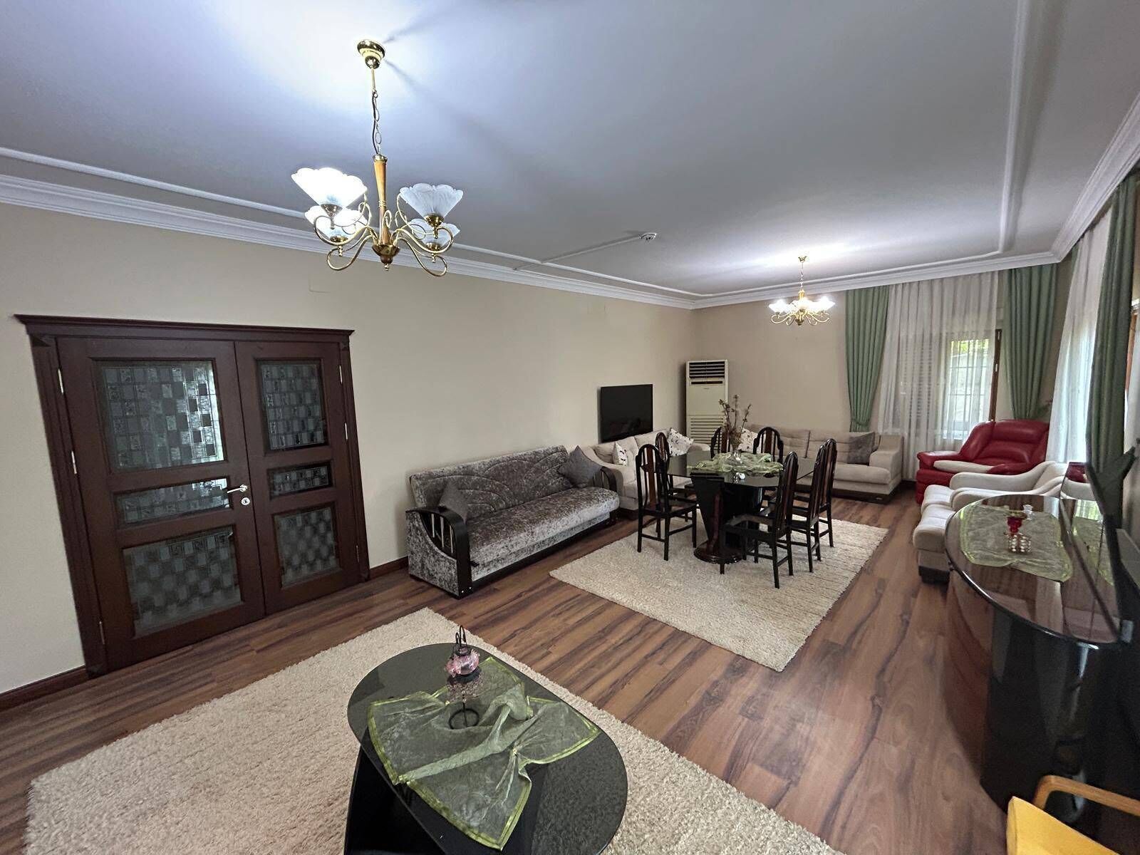A living room with a sofa, dining table, chairs, a chandelier, and a large window with curtains.