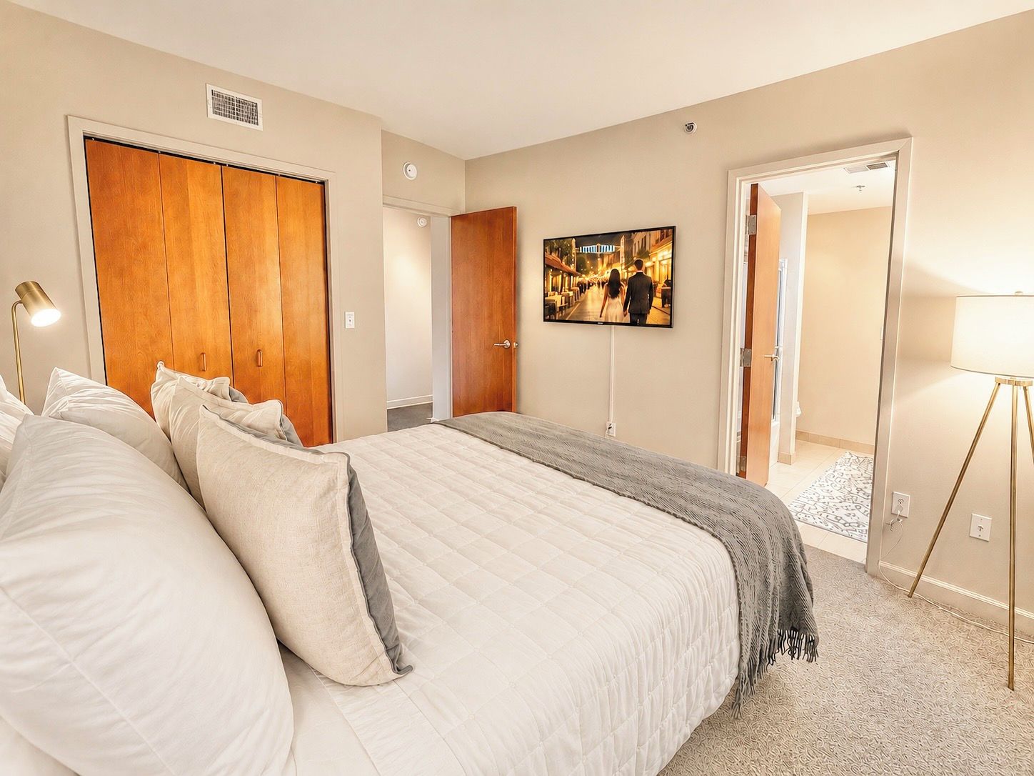 A bedroom with a large bed, a wardrobe, a television on the wall, and a lamp.