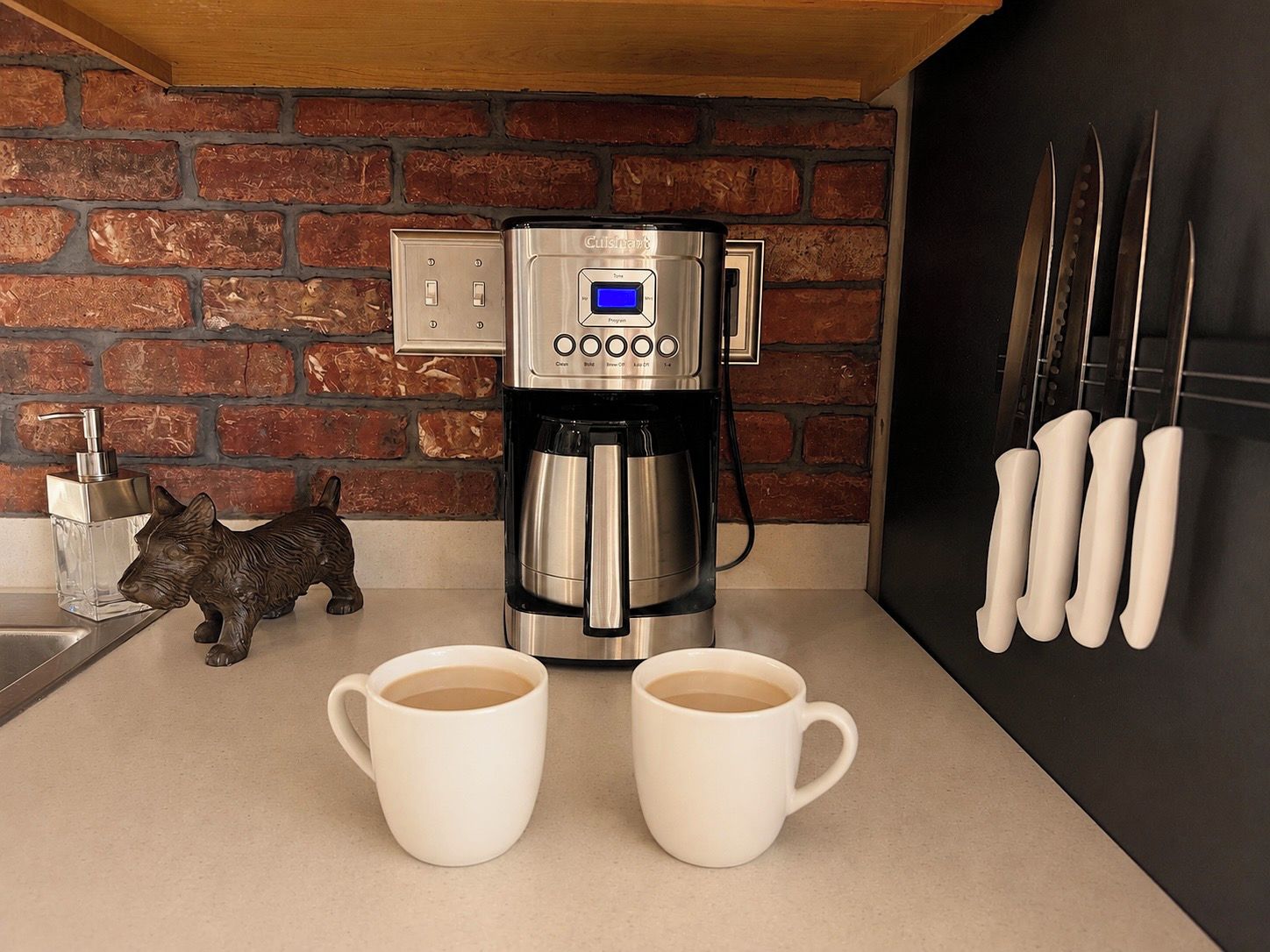 Una cafetera con pantalla digital, dos tazas blancas, un dispensador de jabón y una figurita de perro en una encimera de cocina.