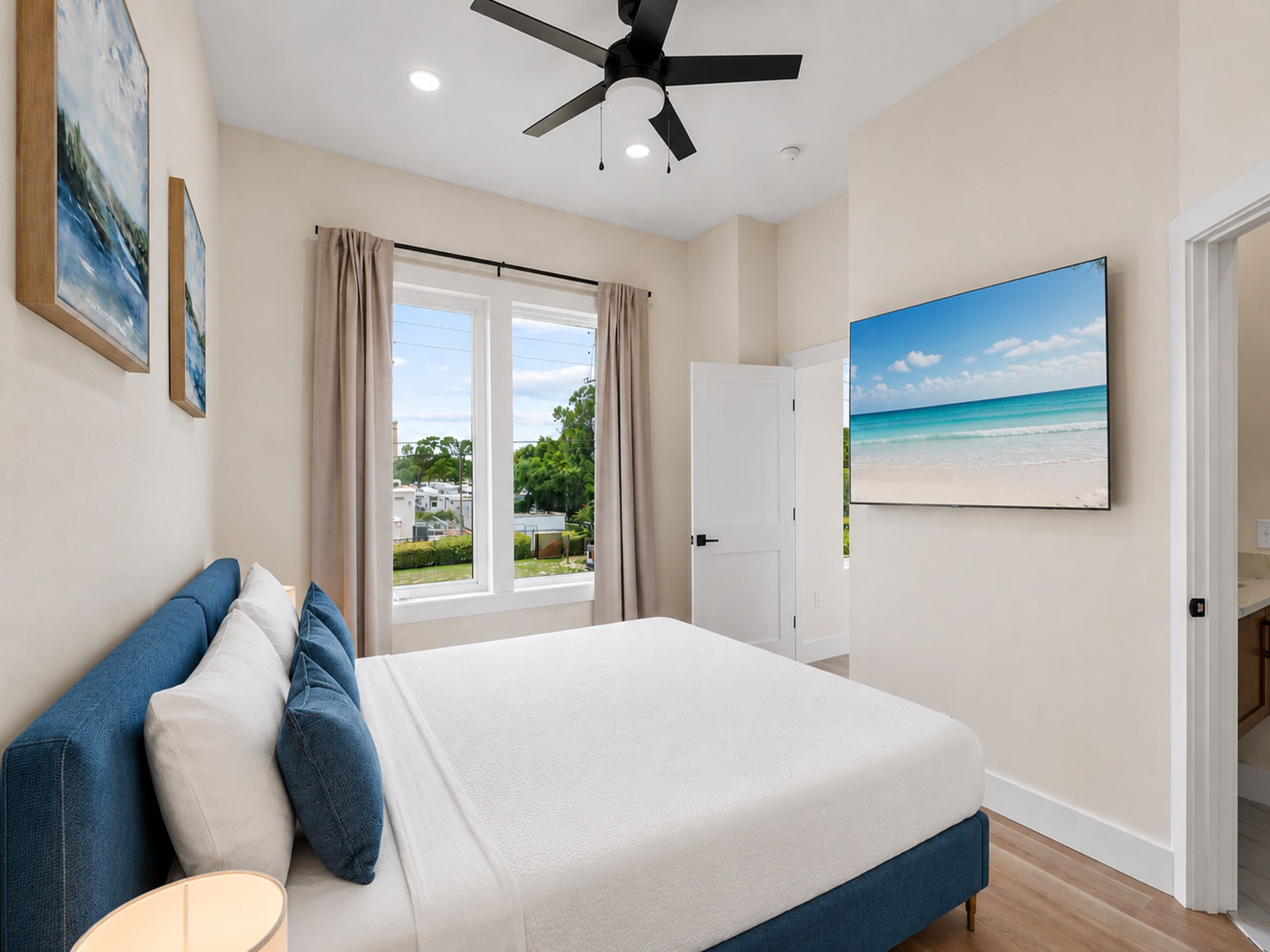A bedroom with a bed, a ceiling fan, a window with curtains, a door, and a beach view painting.