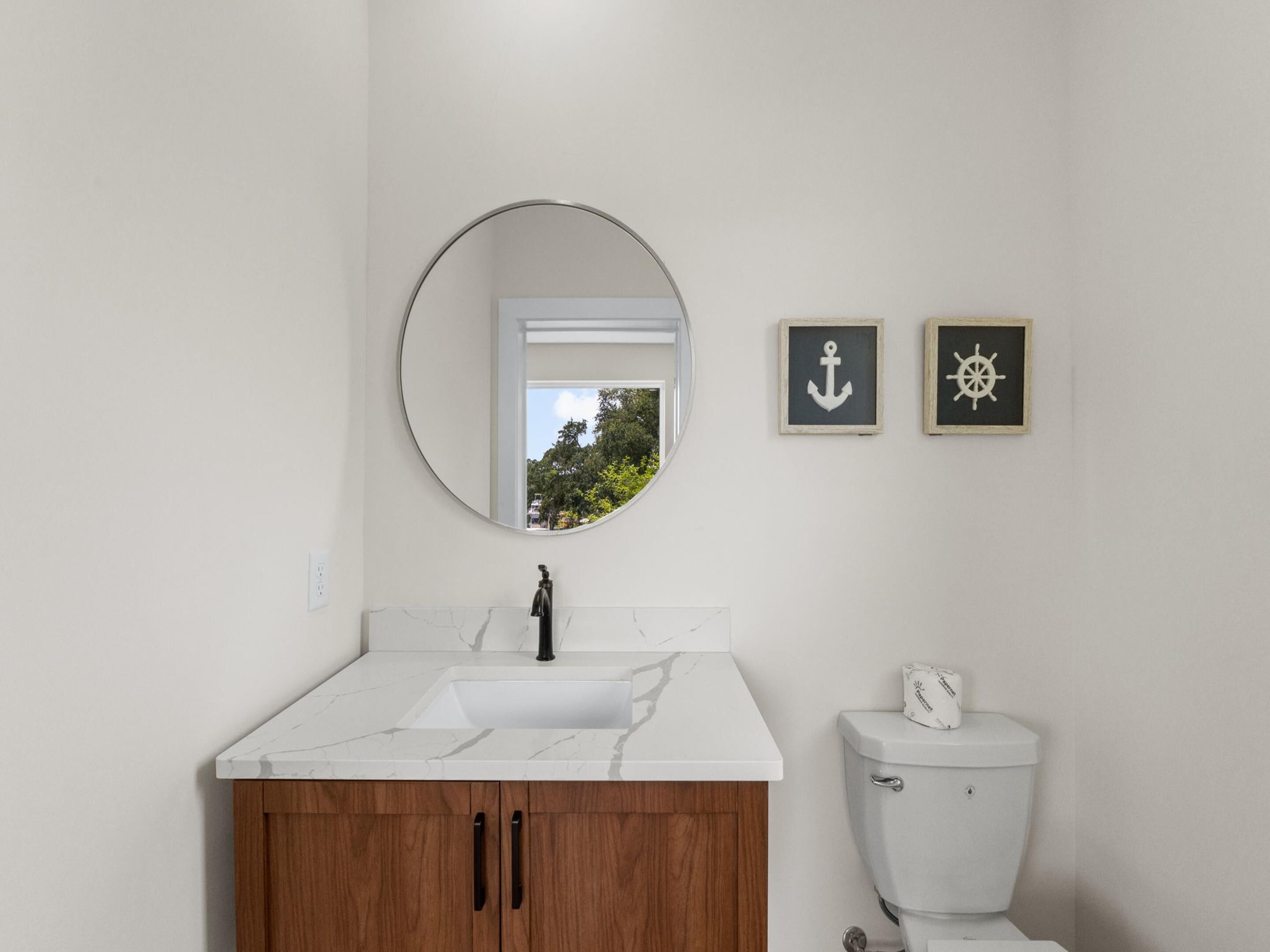 Un baño con un lavamanos blanco, armarios de madera, un inodoro, un espejo redondo y dos cuadros enmarcados con motivos náuticos.