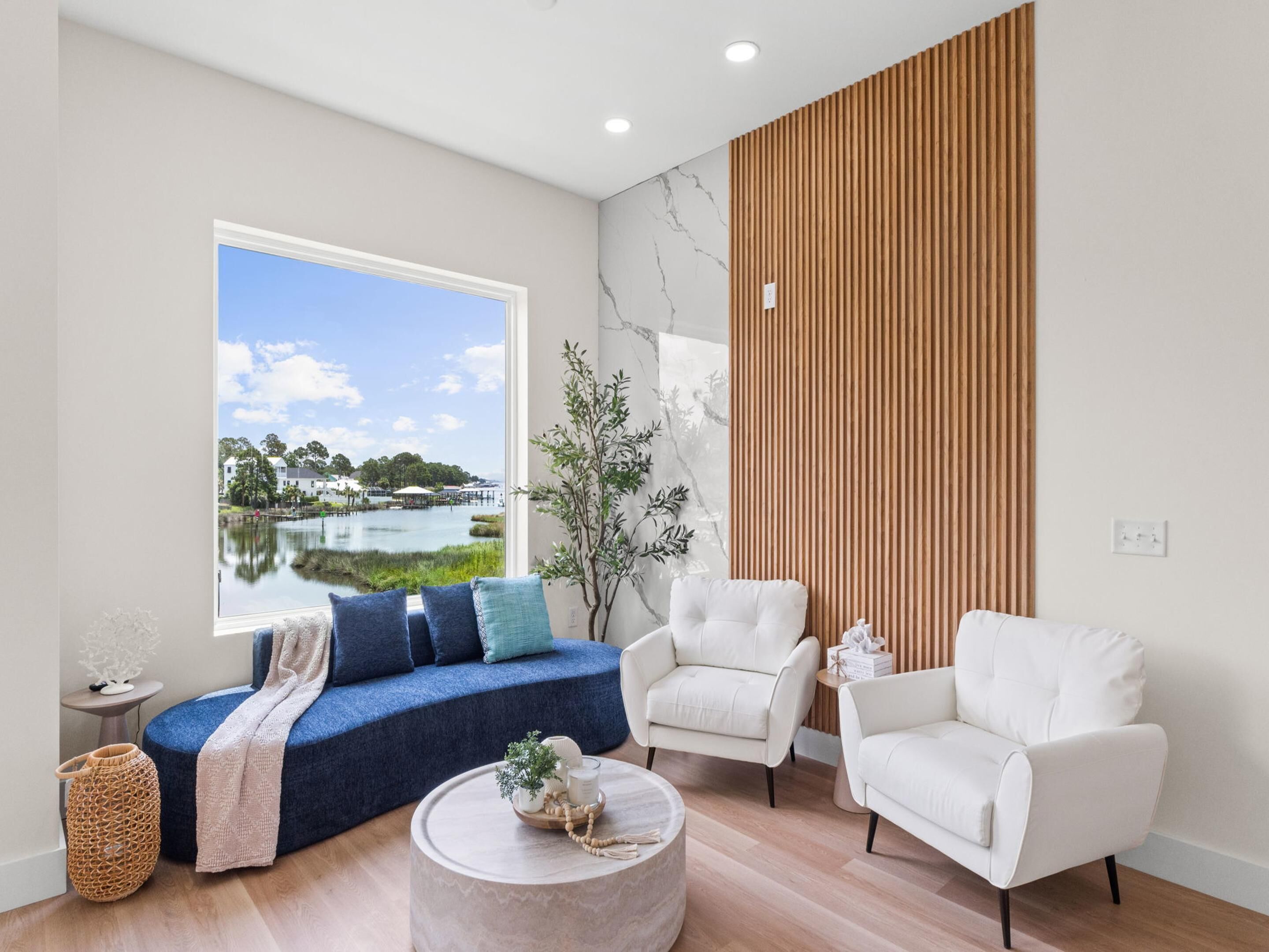 Uma sala de estar moderna com uma janela ampla, um sofá azul, duas poltronas brancas, uma mesa de centro redonda e um painel de ripas de madeira.