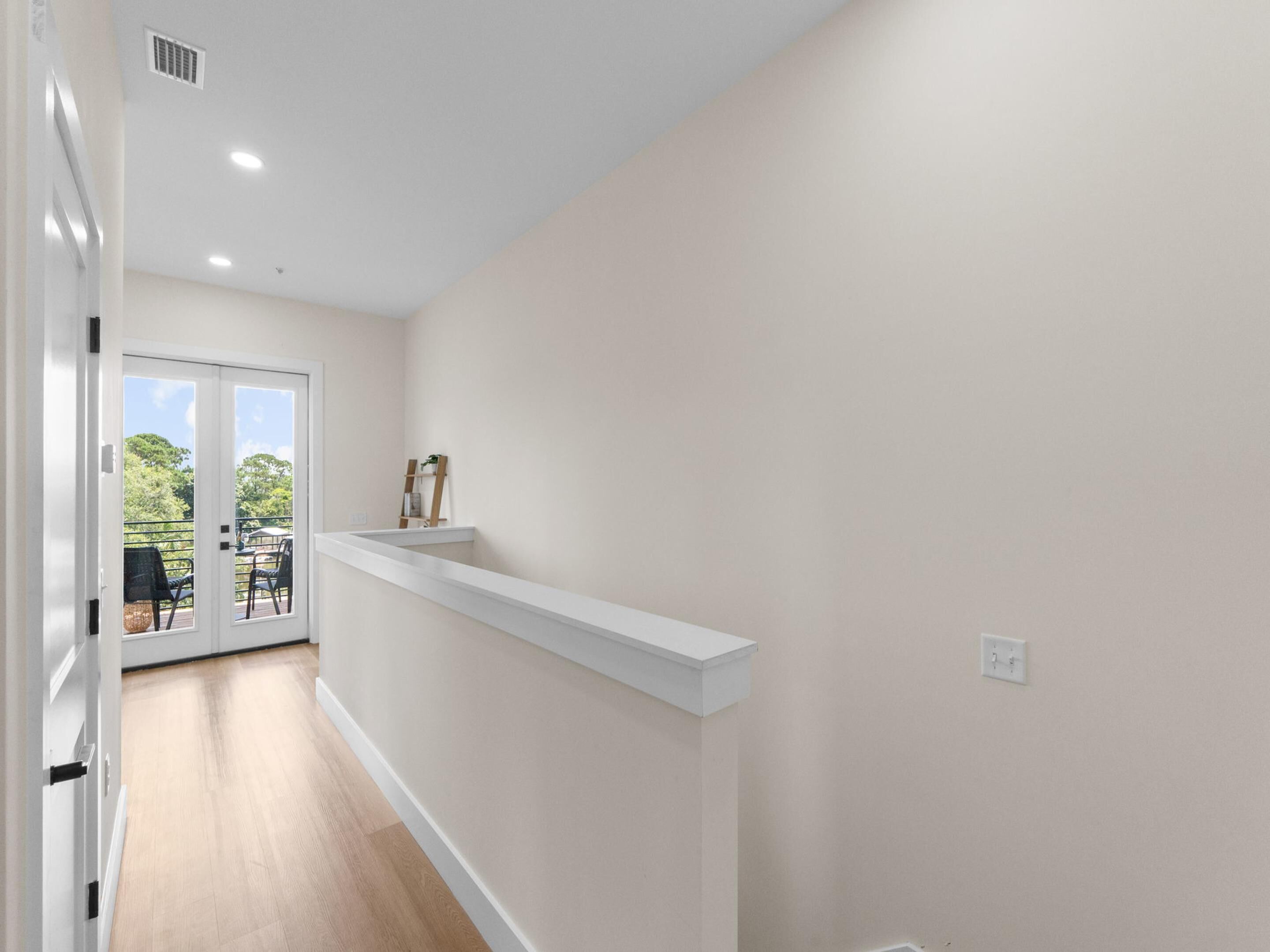 A hallway with a white wall, a wooden floor, and a door leading to an outdoor area with chairs and a table.