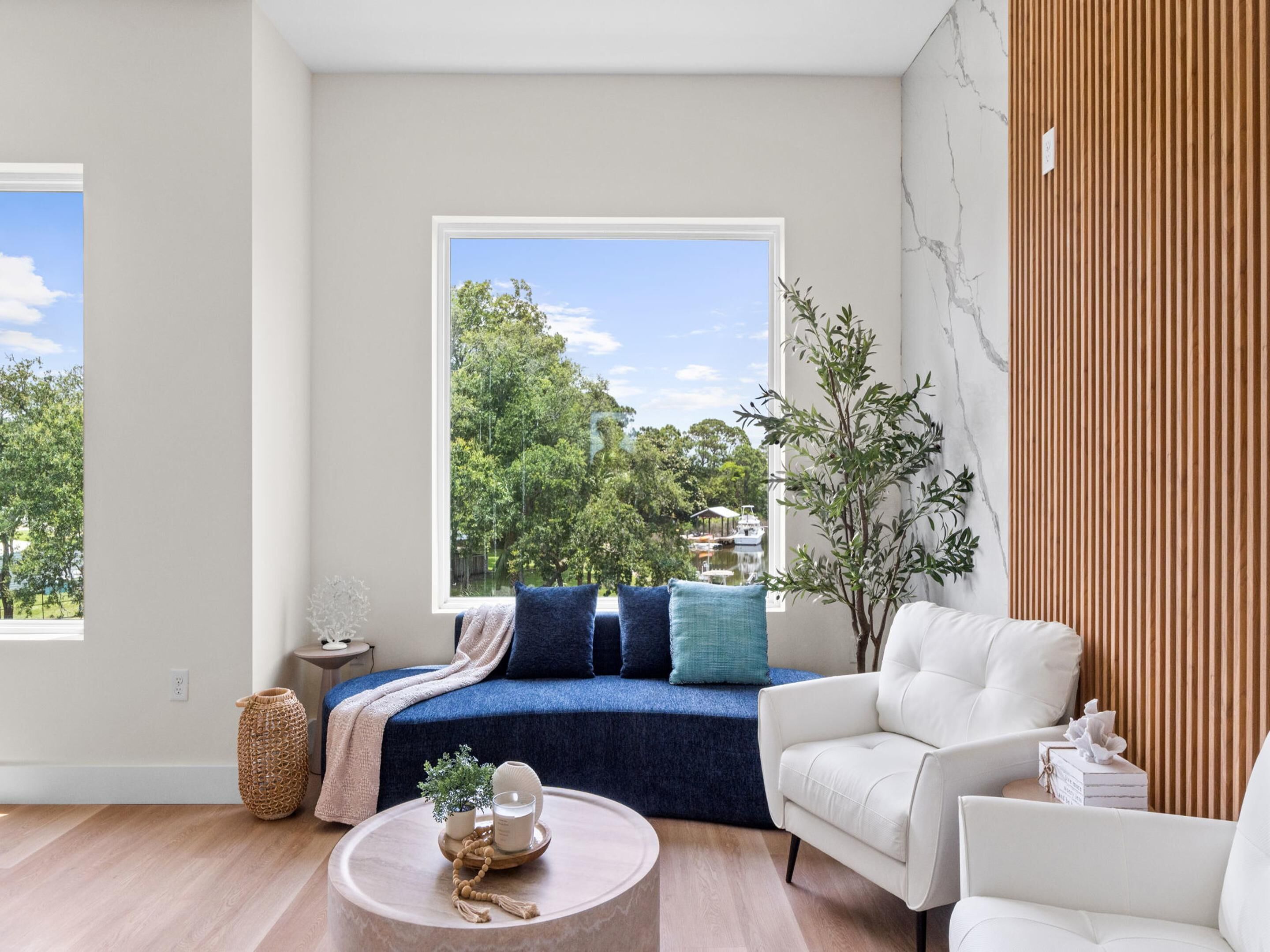 A modern living room with a large window, a blue sofa, a white armchair, and a round coffee table.