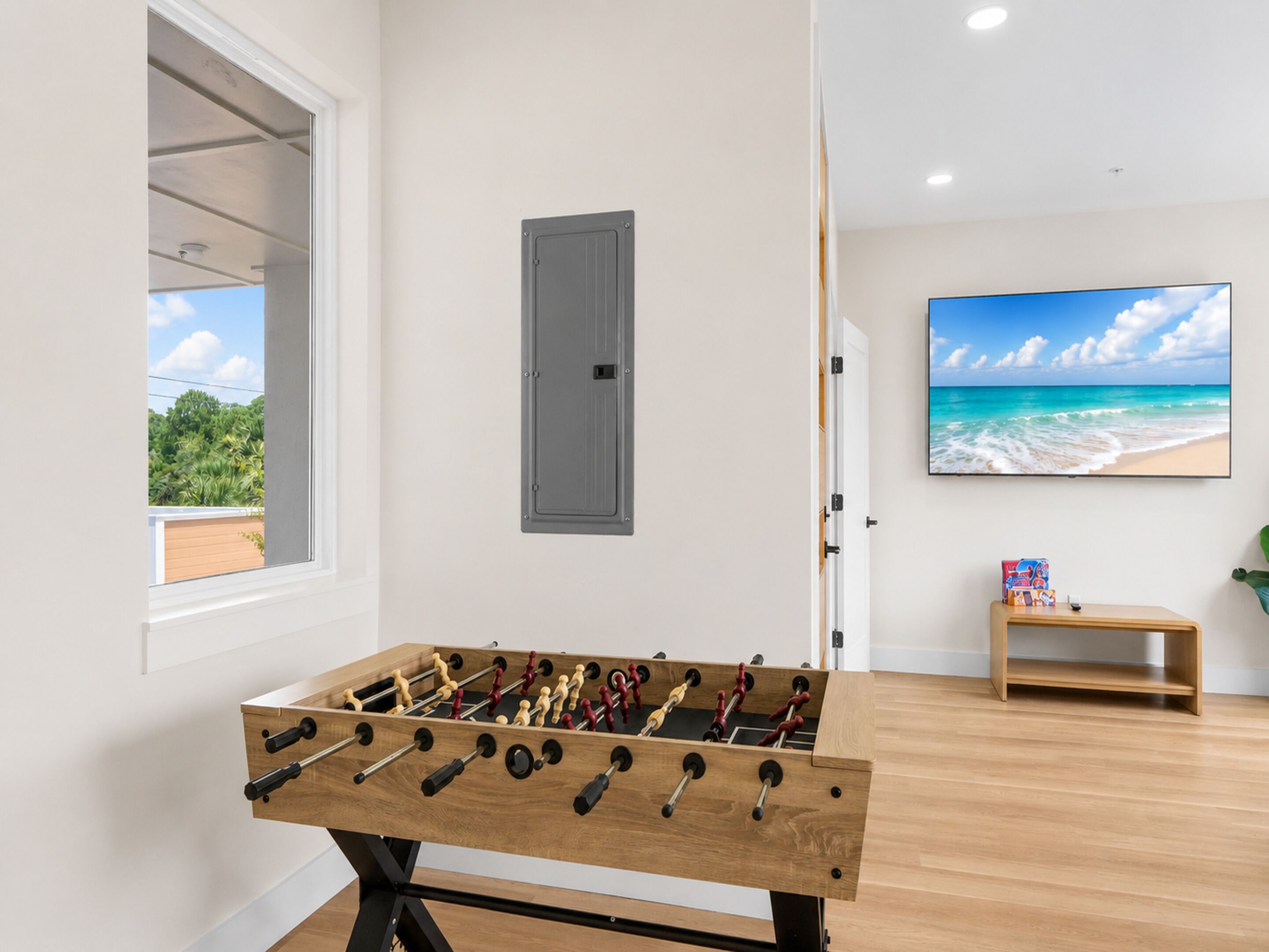 Une pièce avec une table de baby-foot, une fenêtre donnant sur un paysage verdoyant, une porte grise et un téléviseur fixé au mur qui diffuse une scène de plage.