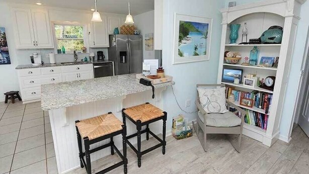 A kitchen with white cabinets, a granite countertop, two stools, a bookshelf, and a painting of a tropical scene. - Paradise Point 3 BR home overlooking the bay in gorgeous Saint George Island (Saint George Island)