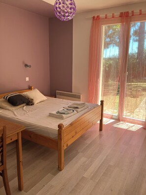 Ein Schlafzimmer mit einem hölzernen Bett, einem Nachttisch, einer Pendelleuchte und Blick durch ein Fenster mit durchsichtigen Vorhängen.