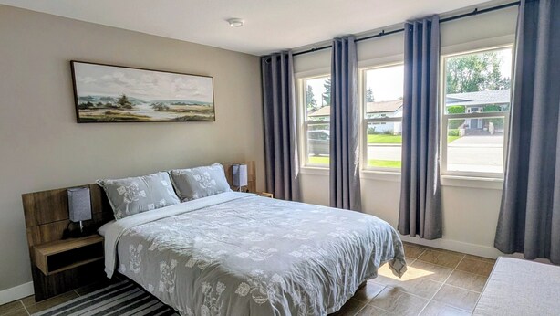Une chambre à coucher avec un lit, une table de chevet, une lampe, un tableau au mur et une vue sur l’extérieur grâce à de grandes fenêtres.
