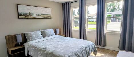 Une chambre à coucher avec un lit, une table de chevet, une lampe, un tableau au mur et une vue sur l’extérieur grâce à de grandes fenêtres.
