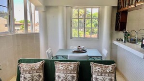 Eine kleine, helle Küche mit Essbereich, ein grünes Sofa mit gemusterten Kissen und ein Fenster mit Blick auf Grünflächen.