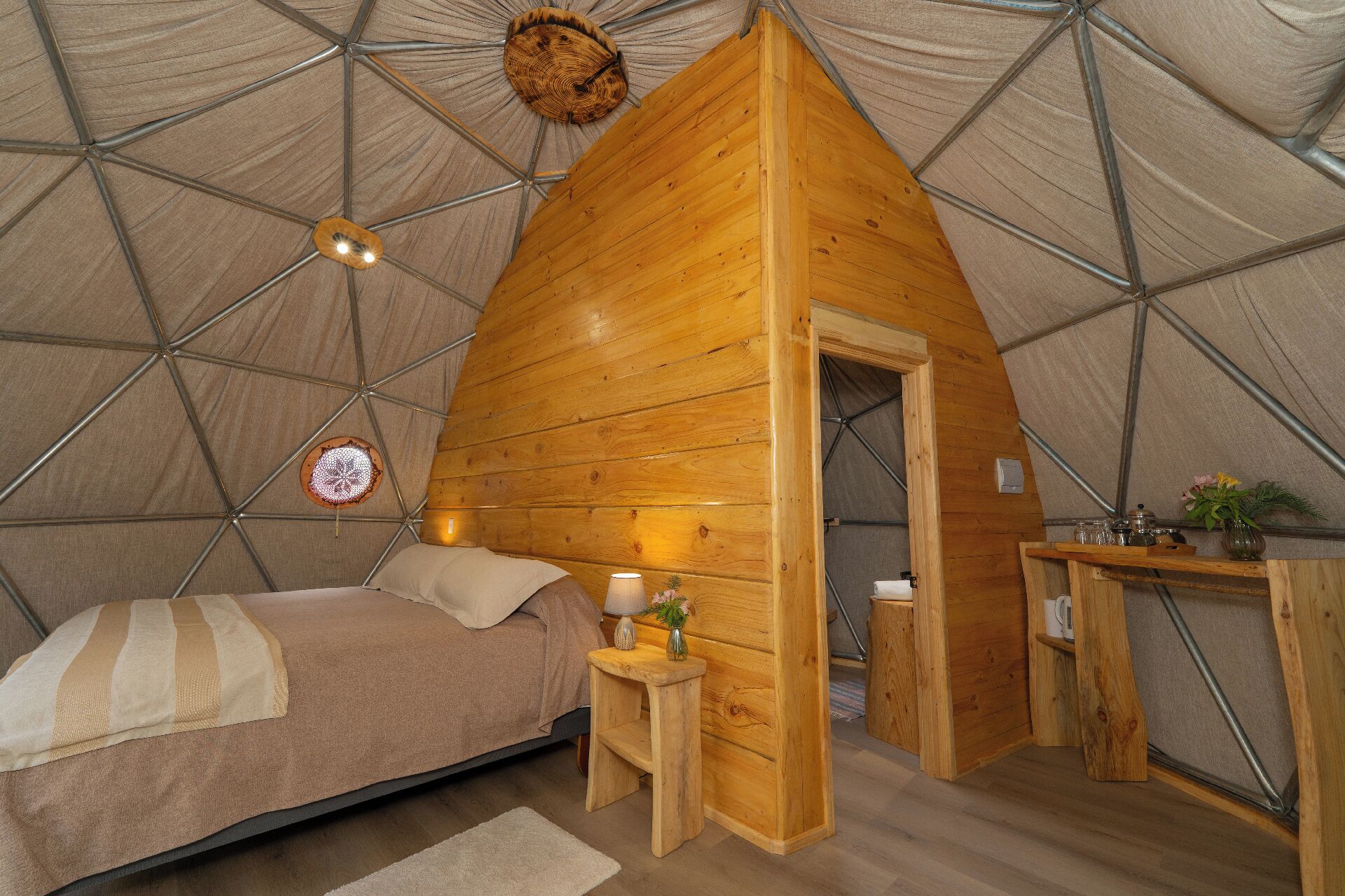 A cozy, triangular wooden tent with a bed, bedside table, lamp, and a small desk with a plant.
