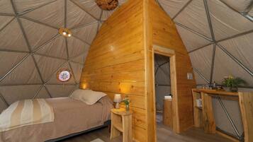 A cozy, triangular wooden tent with a bed, bedside table, lamp, and a small desk with a plant.