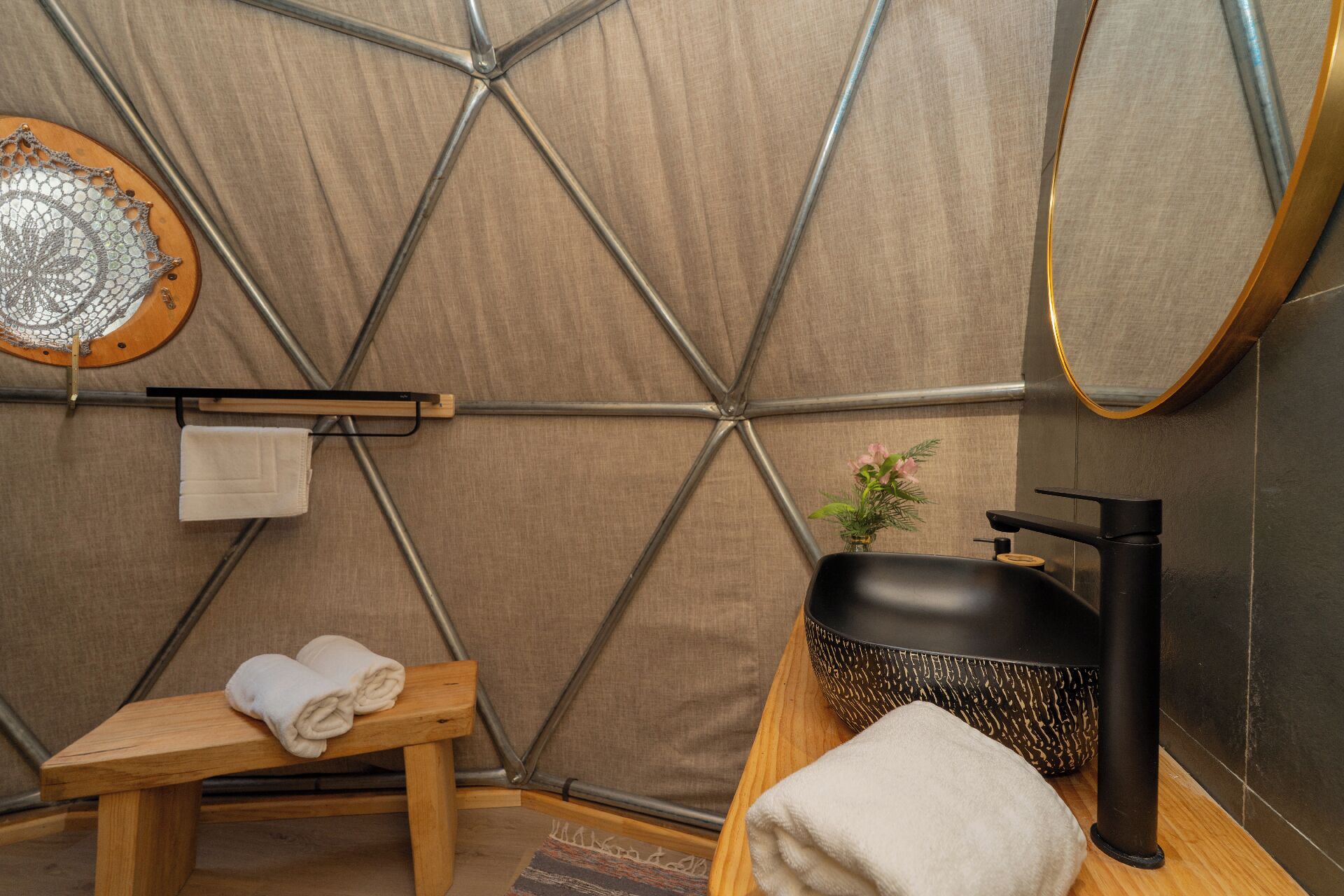 Una habitación tipo tienda de campaña con un banco de madera, una toalla, un lavabo con lavamanos negro y un espejo.