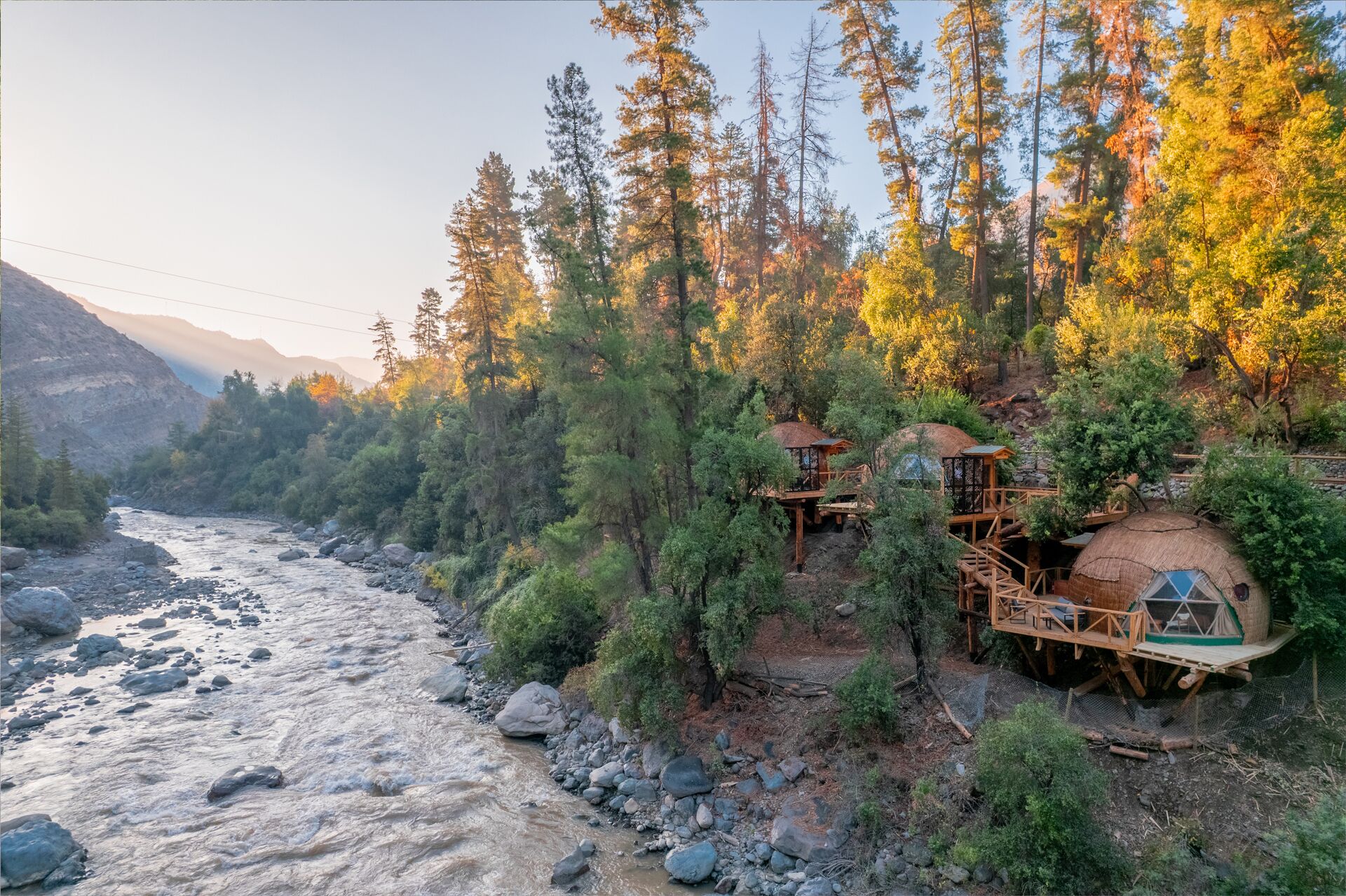 A riverside lodge with multiple treehouses connected by wooden walkways, surrounded by dense forest and mountains.
