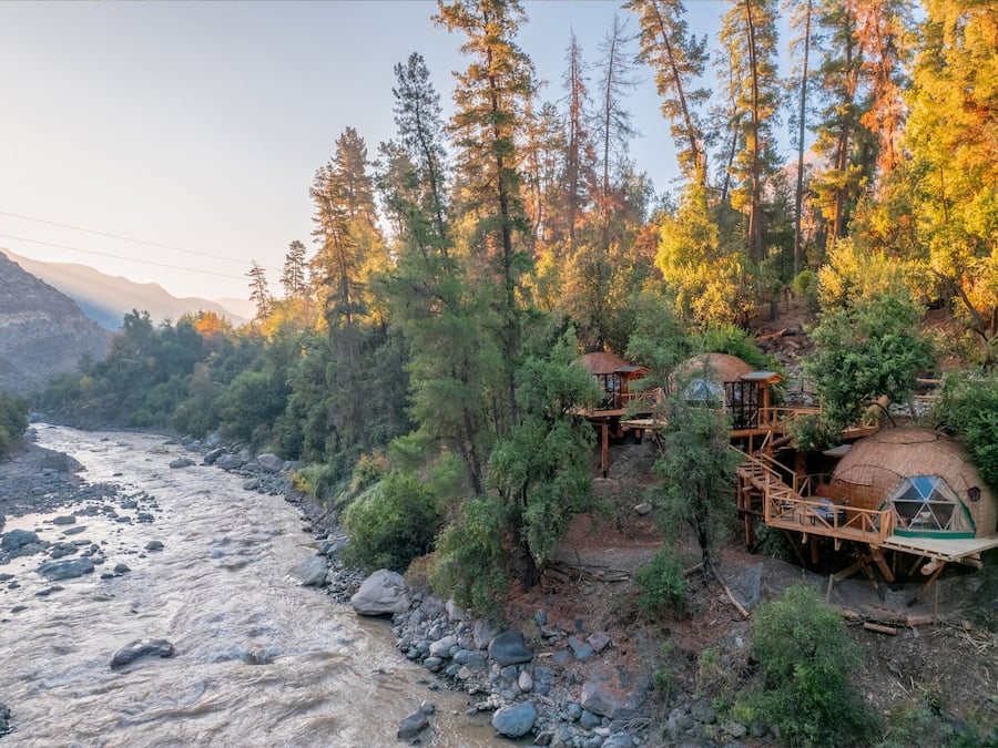 Un chalet au bord de la rivière, doté de plusieurs cabanes dans les arbres reliées par des passerelles en bois, et entouré d’une forêt dense et de montagnes.